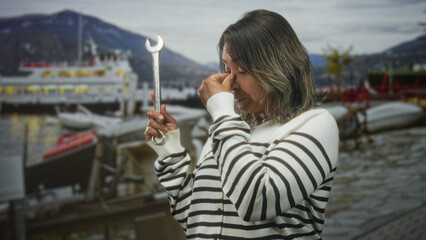 Fototapeta premium Hispanic woman mechanic holding wrench pinching nose on street by marina with ferry and boats; fatigue concern.