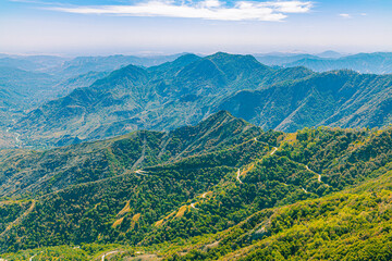 A vast mountain landscape featuring layers of blue ridges and a winding road descending into the valley under a hazy summer sky. © ZL Visuals