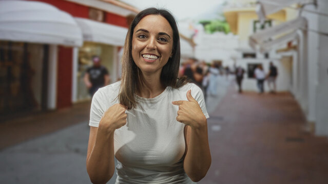 Woman in white t shirt smiling, points thumbs to chest, rings visible, standing on a busy pedestrian street with awnings and storefronts; self confidence.