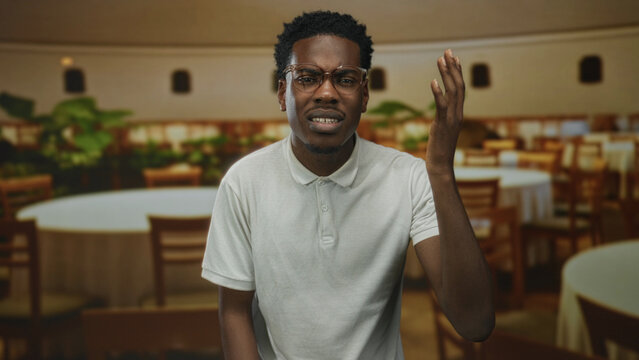 African american man wearing white polo shirt raising right hand gesture inside restaurant building; confusion.