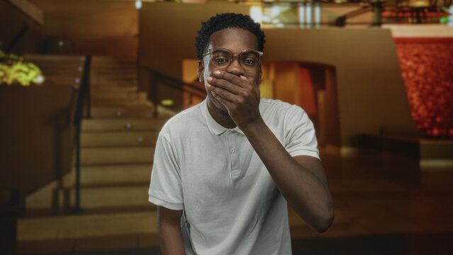 Young african american man wearing white polo shirt and eyeglasses covers mouth with hand by grand staircase in hotel building; surprise.