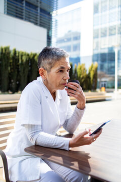 Senior female healthcare worker drinking coffee and using smartphone outdoors