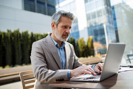 Senior businessman working on laptop at outdoor cafe in modern city