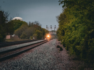Approaching locomotive with bright headlights on double tracks passing under a signal bridge and near a water tower in a suburban forest preserve © Donald