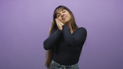 Fototapeta premium Woman resting hands on cheek in studio with purple backdrop and closed eyes and slight smile; calm rest.