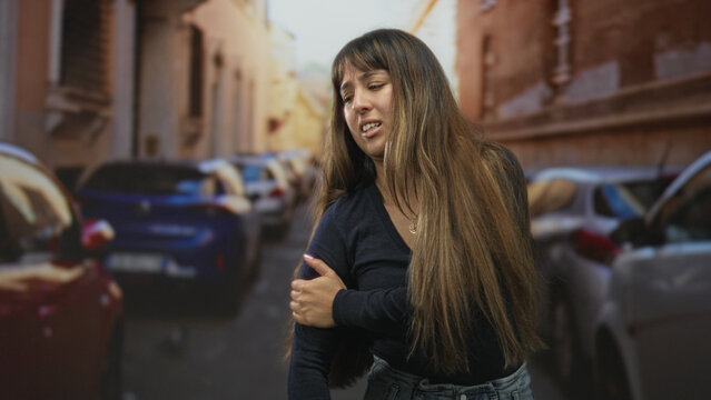 Woman clutching her shoulder with bare hand on a narrow urban street, long hair, eyes closed and winced face amid parked cars; discomfort.