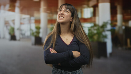Fototapeta premium Woman wearing glasses with arms crossed, smiling and leaning slightly forward in a busy airport terminal with tall columns and planters; confidence approachability warmth.