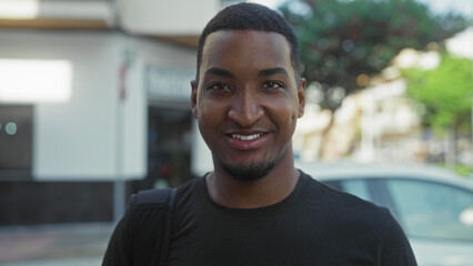 Fototapeta premium Young black man smiling with visible teeth and backpack strap, facing camera on street near parked car and storefront; approachability confidence.
