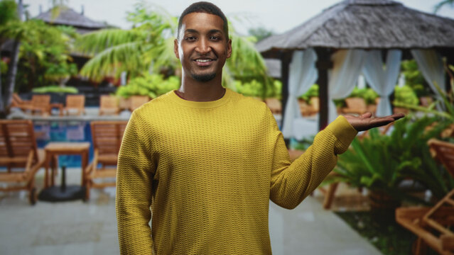 Man presenting palm up at building pool cabana wearing yellow sweater, palm extended in gesture of offering; welcoming calm.