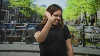 Fototapeta premium Young woman waves hand and smiles wearing black t shirt on amsterdam street near canal and parked bicycles; happiness.