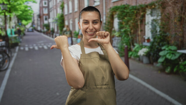 Woman in olive apron pointing thumbs to chest while smiling on a narrow brick street with bicycles and potted plants; pride.