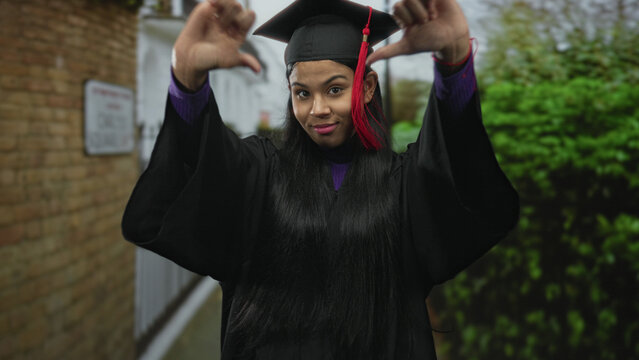 Woman in graduation cap and gown giving thumbsdown gesture on street near building facade; rejection.