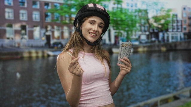 Woman wearing helmet and pink tanktop holding fanned hundred dollar bills and making a finger heart gesture on a street canal in amsterdam; playful wealth tease.