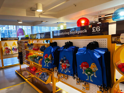 Colorful M and M candy merchandise display with blue drawstring backpacks and gift items in a bright London UK store interior showing sweets, souvenirs and playful retail shelving