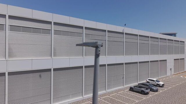 Modern large gray warehouse building facade with horizontal louvre panels, roller doors and parking lot against clear blue sky
