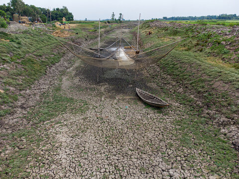 Aerial view of a dried-up riverbed with cracked earth, traditional fishing nets on bamboo poles, a small wooden boat, and people on the banks in Natore, Rajshahi Division, Bangladesh.