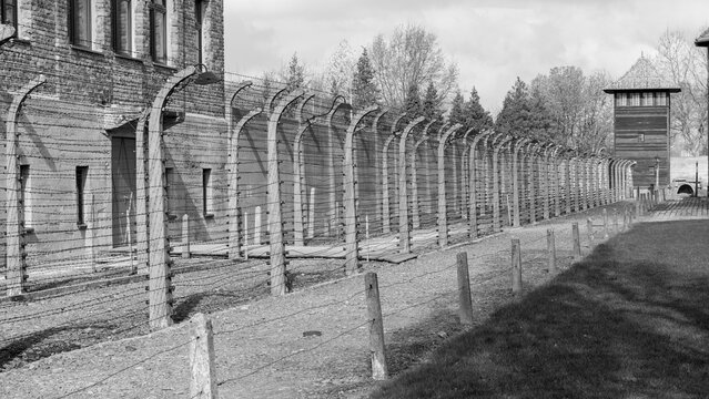 Black and white perspective of electrified barbed wire fences and watchtower at Auschwitz I. Grim view of the perimeter fortification at the Holocaust memorial and museum in Oswiecim, Poland.
