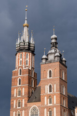 Fototapeta premium The asymmetric towers of St. Mary's Basilica in Krakow. A detailed view of the Gothic brick church's spires against a dark, stormy sky in the Main Market Square, Poland.