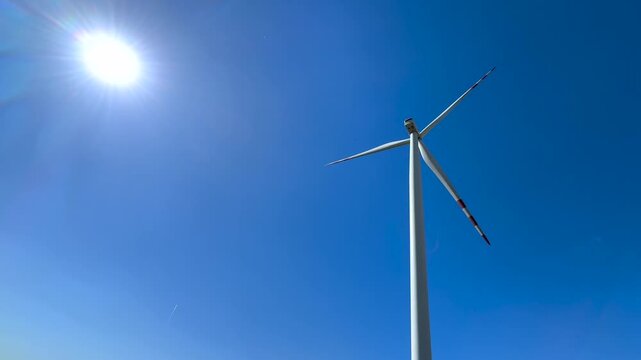 Low angle view of a modern wind turbine rotating its large blades against a bright sun and a clear blue sky, generating clean, renewable energy for a sustainable future and innovation