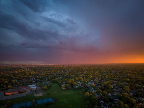 Wide aerial drone view of a dramatic orange sunset and dark storm clouds over a lush green suburban neighborhood in Naperville Illinois