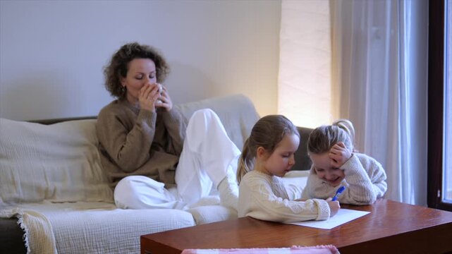 Cozy family evening at home where a mother enjoys a hot drink relaxing on the couch while her two young daughters are happily drawing together at a coffee table in the living room