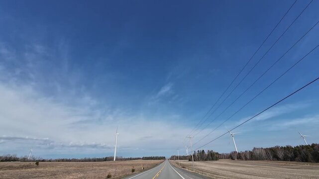 4K dashcam footage driving on a rural road lined with wooden power poles and wind turbines in yellow spring fields near Baie-des-Sables, Quebec, Canada.