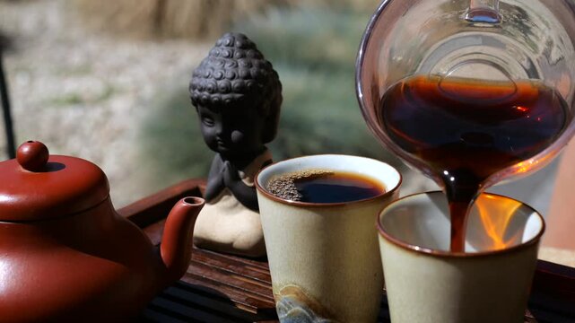 Pouring dark pu erh tea from a glass serving pitcher into small ceramic cups during a traditional gongfu cha tea ceremony with a buddha statuette and yixing teapot on a wooden tray