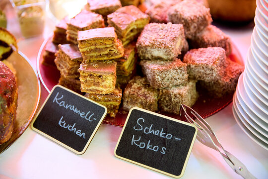 Assorted cake slices on a platter at a buffet, featuring caramel and chocolate-coconut flavors with handwritten chalkboard labels.
