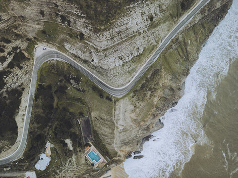 Aerial view of rugged cliffs and winding coastal road beside the blue ocean with white waves, Buarcos e Sao Juliao, Coimbra, Portugal.