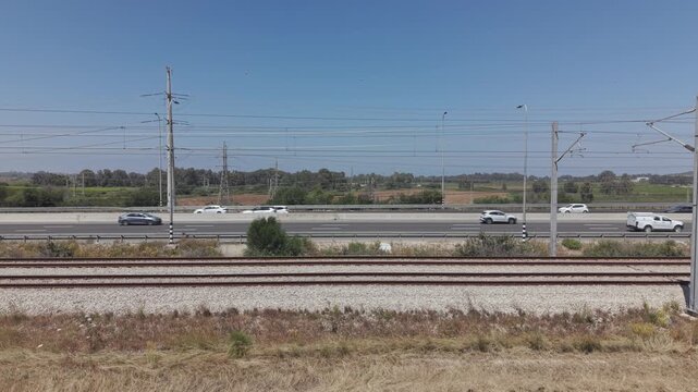 Wide view of railway track in foreground and highway with traffic and power lines across flat landscape in Israel
