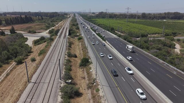 Aerial drone view of multilane highway running parallel to railway tracks with traffic through green landscape in Israel

