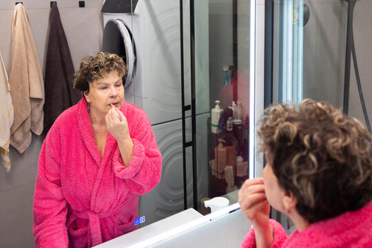Senior woman standing in front of mirror moisturizing lips with balm, morning hygiene routine and healthy lifestyle concept.