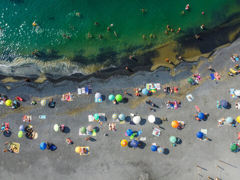 Aerial view of people sunbathing under colorful umbrellas on a pebble beach and swimming in turquoise water in San Lorenzo, Liguria, Italy.