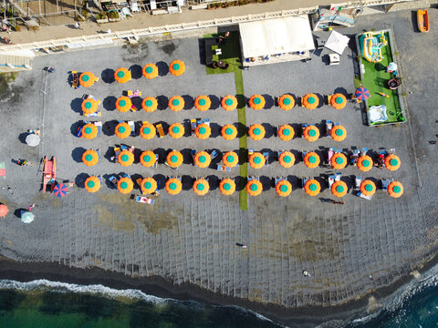 Aerial view of organized rows of colorful beach umbrellas on a pebble shore with clear turquoise water in Imperia, Liguria, Italy.
