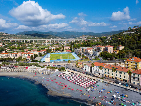 Aerial view of the sandy coastline with beach umbrellas, a sports stadium, and a large highway viaduct against green hills in Imperia, Liguria, Italy.