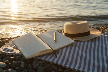 Open notebook with pen resting on striped beach blanket near calm sea water at golden sunset, straw hat beside journal creating peaceful summer vacation atmosphere, travel lifestyle concept © Evgeniia Primavera