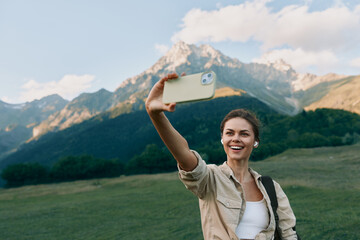 Fototapeta premium A cheerful woman takes a selfie in a scenic mountain meadow, casual jacket, sunny day. Lifestyle portrait capturing outdoor exploration and joyful travel moments, authentic, bright, modern.