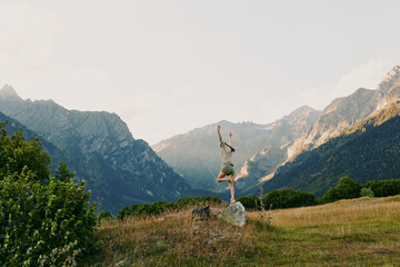 Obraz premium Young woman leaps on a rock in a sunlit alpine meadow with distant rugged mountains, radiating freedom and joy in an outdoorsy, carefree moment