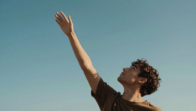 Young man reaches up with his hand towards the light blue sky on a bright day
