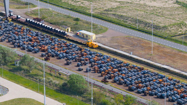 Aerial view of the Tata Steel industrial site featuring rows of large steel coils stored outdoors next to a yellow transport vehicle and a railway track in Velsen-Noord, Noord-Holland, Netherlands.