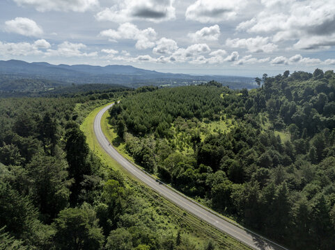 Aerial view of the winding asphalt road passing through the dense Lembus Forest with rolling hills under a cloudy sky in Eldama Ravine, Baringo County, Kenya.