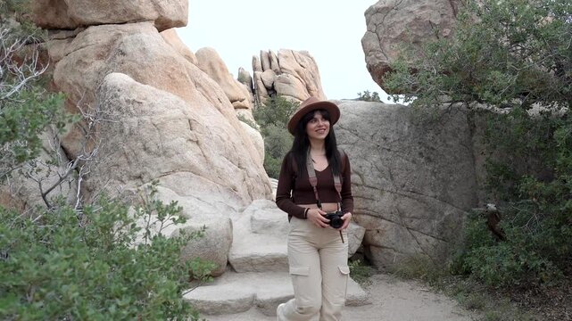 Young smiling woman with a vintage camera walking down stone steps on a hiking trail in hidden valley, exploring the rocky landscape of joshua tree national park in california during her vacation