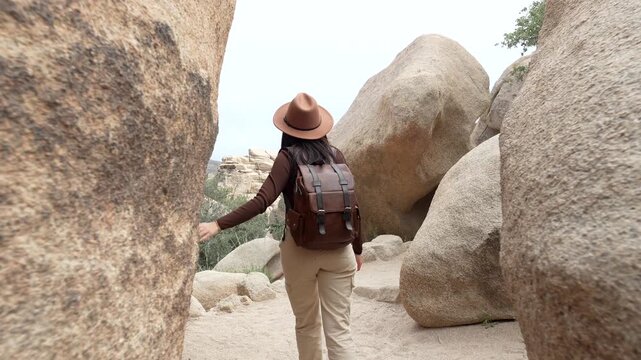 Adventurous young woman with a backpack hiking through a narrow passage between large rock formations on the hidden valley nature trail in joshua tree national park, california