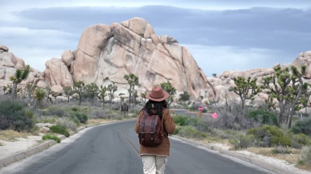 Young female traveler with a backpack and hat walking down an empty road surrounded by yucca trees and giant boulders inside the beautiful joshua tree national park in california