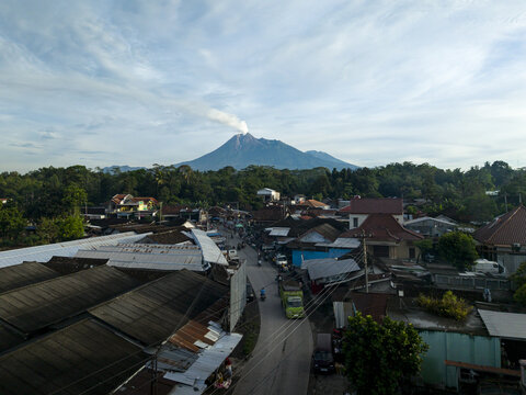 Aerial view of Pasar Kembang Klaten traditional market with rooftops, a winding road, and the smoking Mount Merapi volcano in the distance Klaten, Central Java, Indonesia.