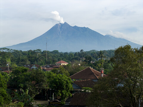 Aerial view of Mount Merapi volcano with smoke rising from the crater above village rooftops and lush tropical greenery in Klaten, Central Java, Indonesia.