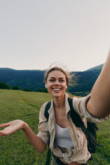 Fototapeta premium Smiling woman with backpack taking a selfie in an open meadow near mountains. Outdoor lifestyle, nature exploration, and travel mood. Bright natural light creates a warm vibe.