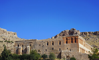 The historic stone facade of the ancient Deyrulzafaran Monastery (Mor Hananyo Monastery) stands at the base of arid, rugged mountains under a clear blue sky near Mardin, southeastern Turkey © Gregory
