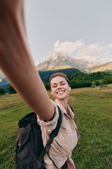 Fototapeta premium Woman outdoors with backpack smiles for a selfie during a hike in a grassy valley beneath mountains, enjoying nature and travel in bright daylight, casual lifestyle