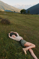 Fototapeta premium Young woman rests on a grassy hillside in a scenic valley, eyes closed and arms stretched overhead, enjoying calm after a hike in bright daylight.